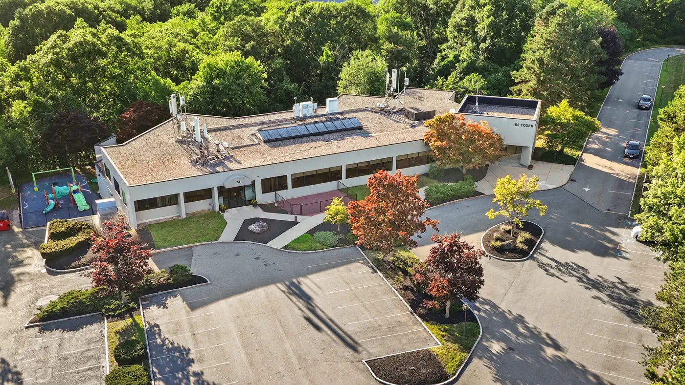 Aerial view of 55 Tozer Road, a two-story standalone office building on a wooded 6-acre campus in Beverly, Massachusetts, with surface parking and a playground